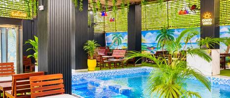 An indoor pool area with wooden chairs, a blue mosaic pool, hanging plants, and a tropical mural.