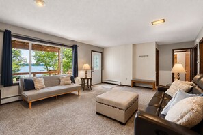 A spacious living room with a sofa, ottoman, and a leather couch, a small table with a lamp, and a view of a lake through large windows. - French Lake Shores-Sunset Retreat/Private 3 Bedroom Lakefront for 8 (Faribault)