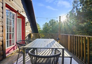 A wooden deck with a table and chairs, a red door, and a view of trees. - Game Loft | Hot Tub | Fire Pit | Wooded Escape (Broken Bow)