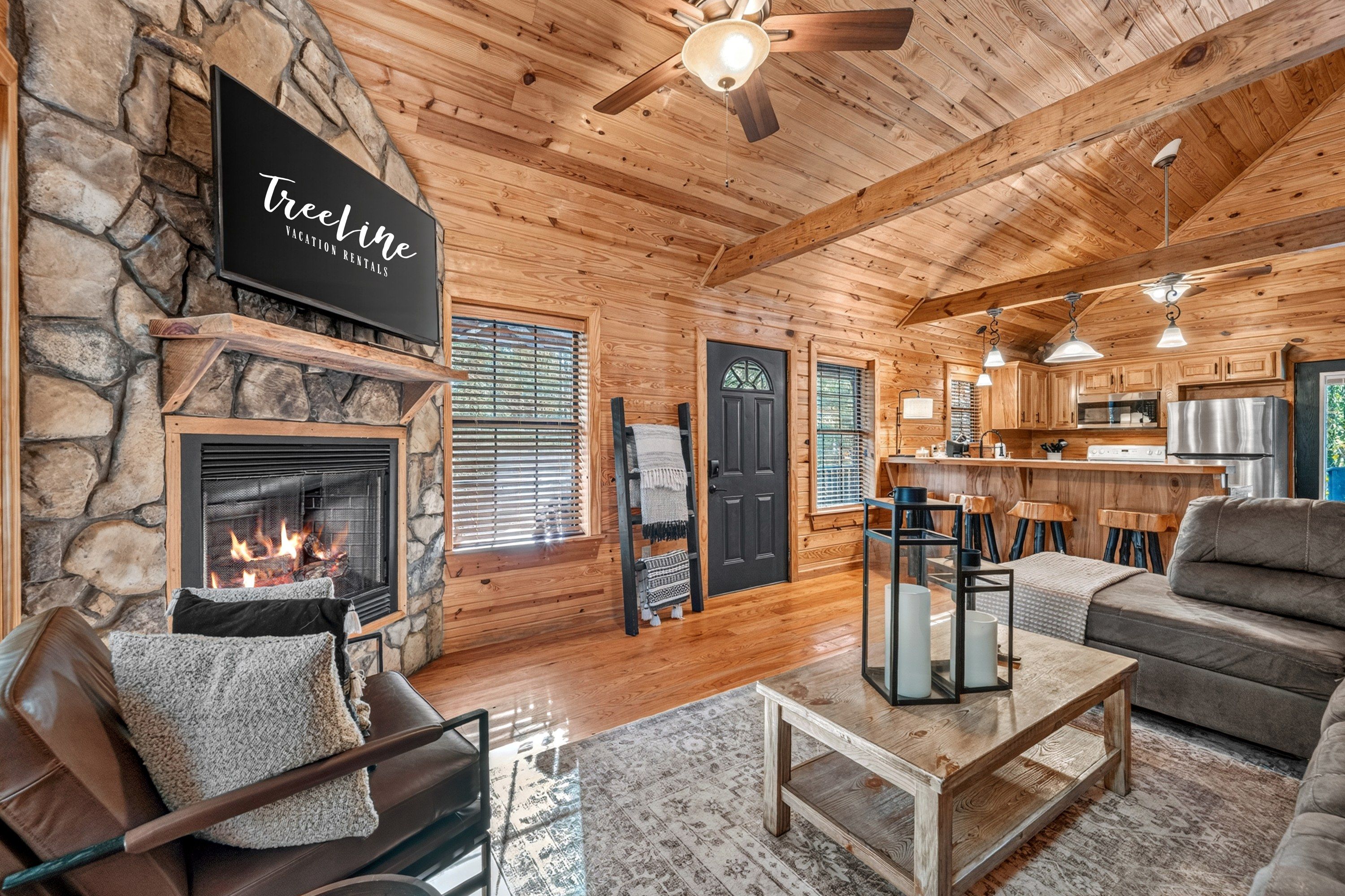 Un intérieur de chalet chaleureux avec une cheminée en pierre, des murs en bois, un coin salon avec un canapé et un fauteuil, et un espace cuisine.