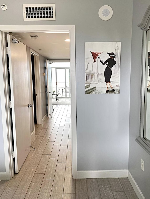 A hallway with a door leading to a balcony, a wall-mounted air vent, and a framed artwork of a woman holding an umbrella. - Renovated oceanfront 1 bedroom condo (Myrtle Beach)