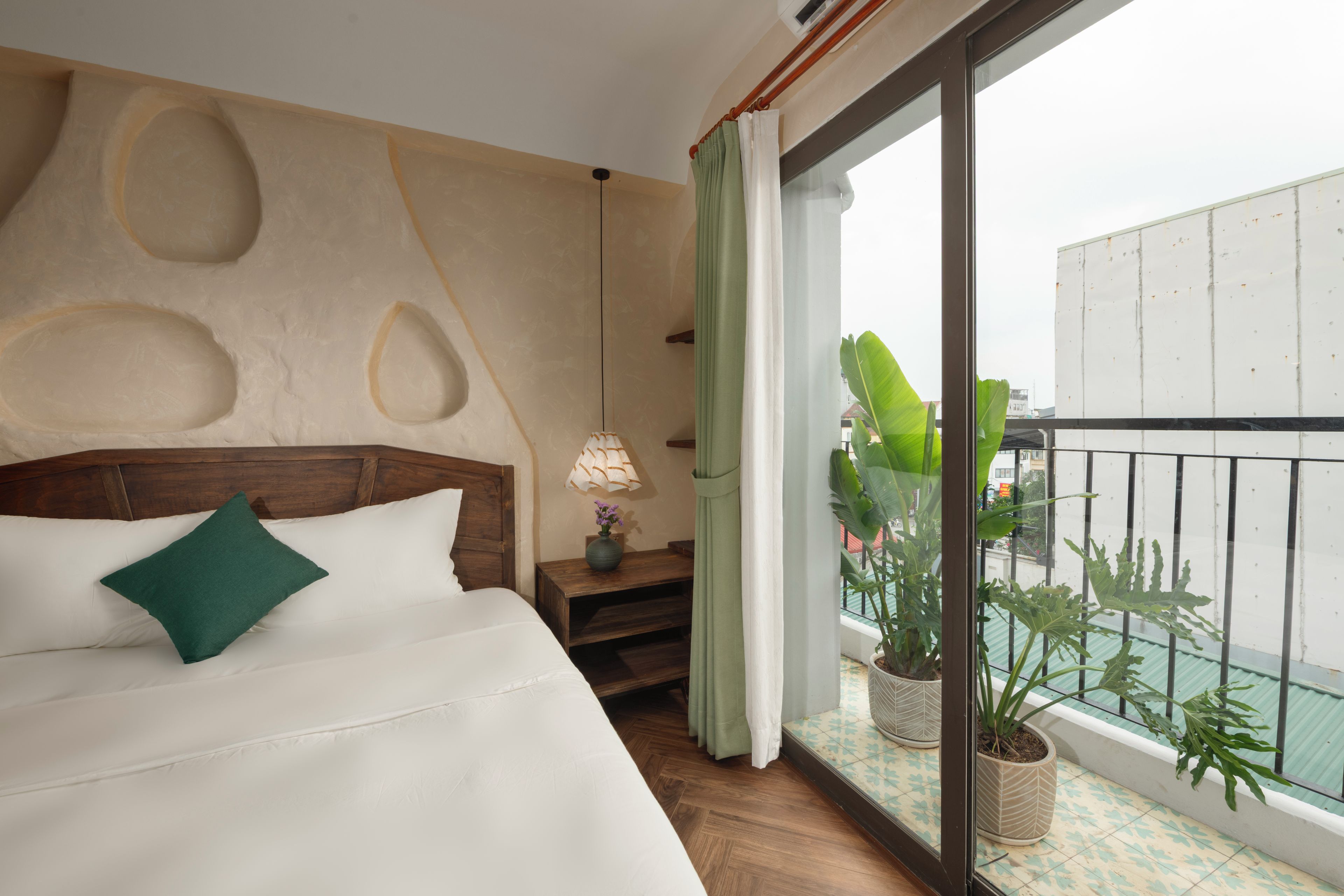 Habitación de hotel con una cama grande, cabecera de madera, mesita de noche con lámpara y vista a la ciudad desde un balcón con plantas en macetas.