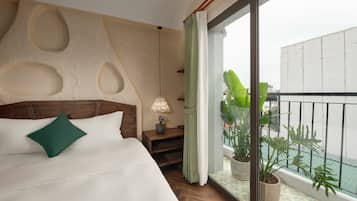 Habitación de hotel con una cama grande, cabecera de madera, mesita de noche con lámpara y vista a la ciudad desde un balcón con plantas en macetas.