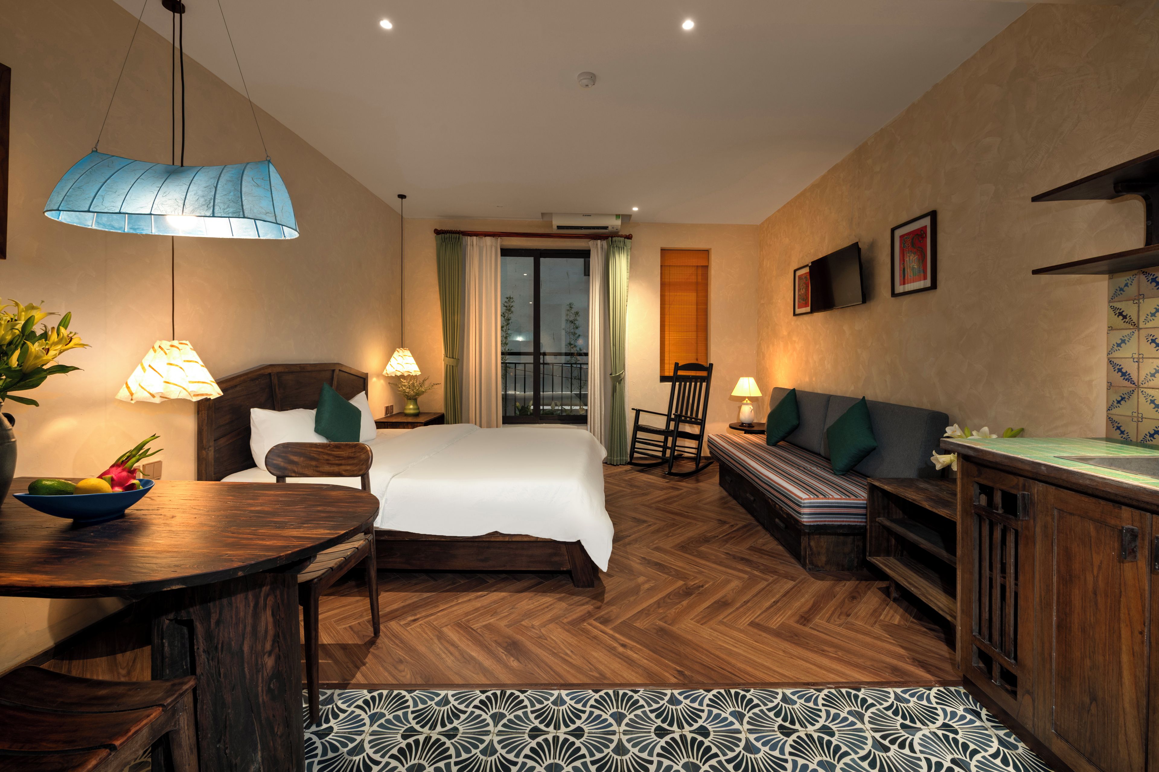 A hotel room with a bed, a seating area with a sofa and chairs, a dining table with a bowl of fruit, and a patterned floor.