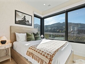 Ein modernes Schlafzimmer mit einem großen Bett, einem Nachttisch, einer Lampe, einem gerahmten Bild und Blick auf eine Stadtlandschaft durch ein großes Fenster.