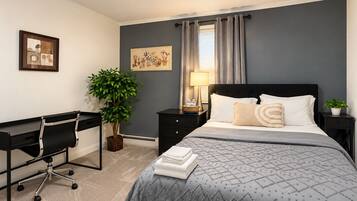 A bedroom with a bed, desk, chair, lamp, and two potted plants.