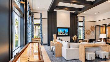 A modern living room with a shuffleboard table, a large flat-screen TV, and a seating area with sofas and ottomans.