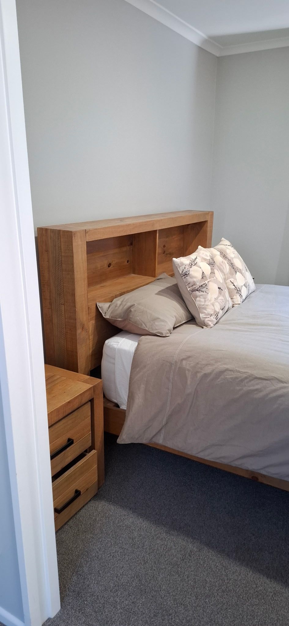 A bedroom with a wooden headboard, a bed with beige bedding, two pillows, and a nightstand with drawers.