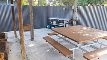Una veranda esterna con tavolo e panche in legno, recinzione in lamiera ondulata e un'area di stoccaggio con contenitori.