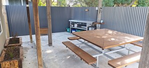 Une terrasse extérieure avec une table et des bancs en bois, une clôture en métal ondulé et un espace de rangement avec des bacs.