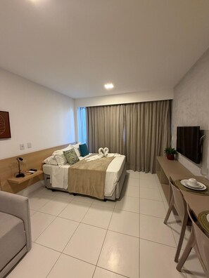 Habitación de hotel con cama, mesita de noche, lámpara, televisión y zona de comedor con mesa y sillas.