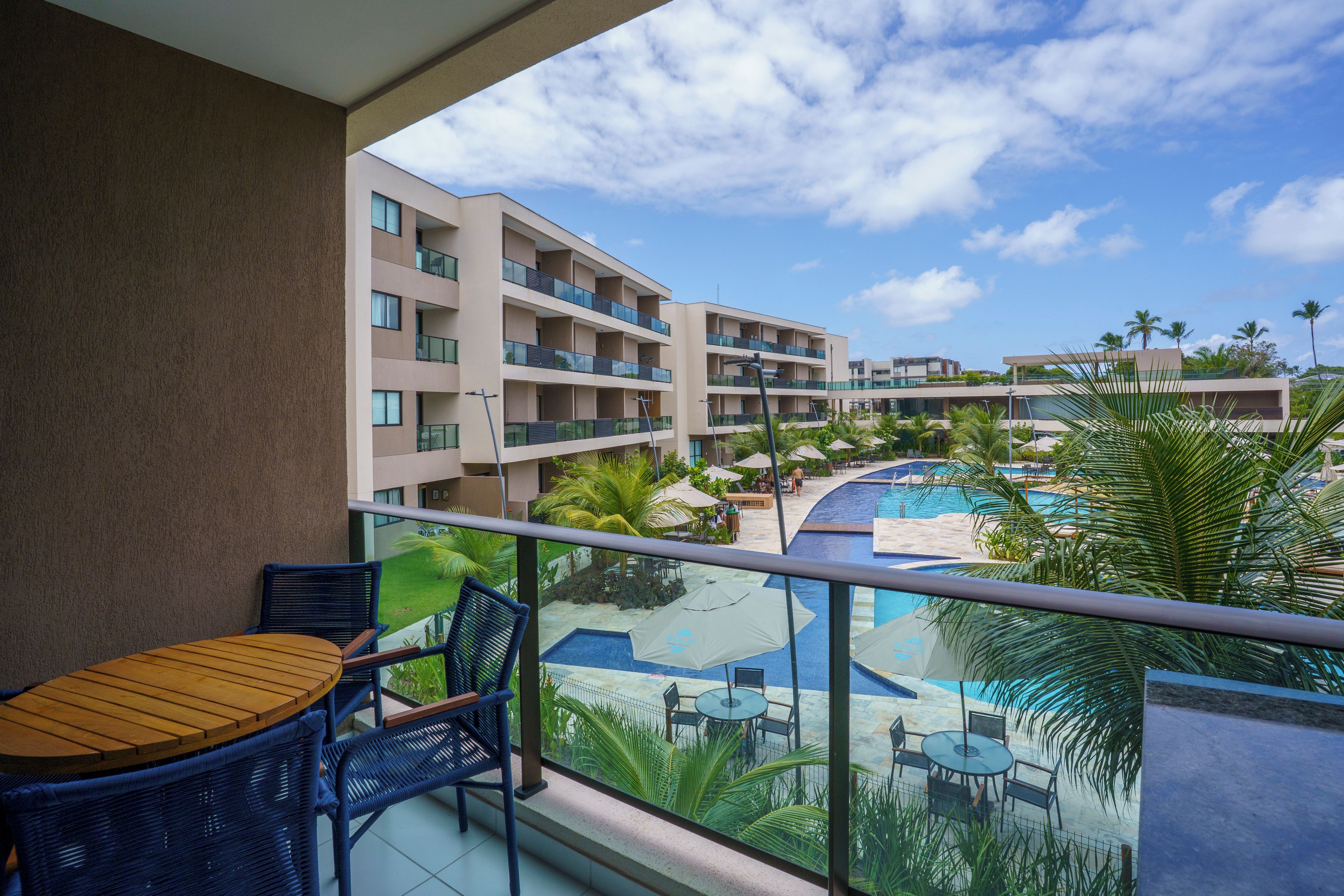 Un balcón con mesa y sillas, con vista a una piscina y un edificio con varios balcones.