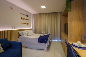 A hotel room with a bed, a blue sofa, a wooden TV stand, and a dining area with a table and chairs. - Malia Beach by Essence Porto de Galinhas (Ipojuca)