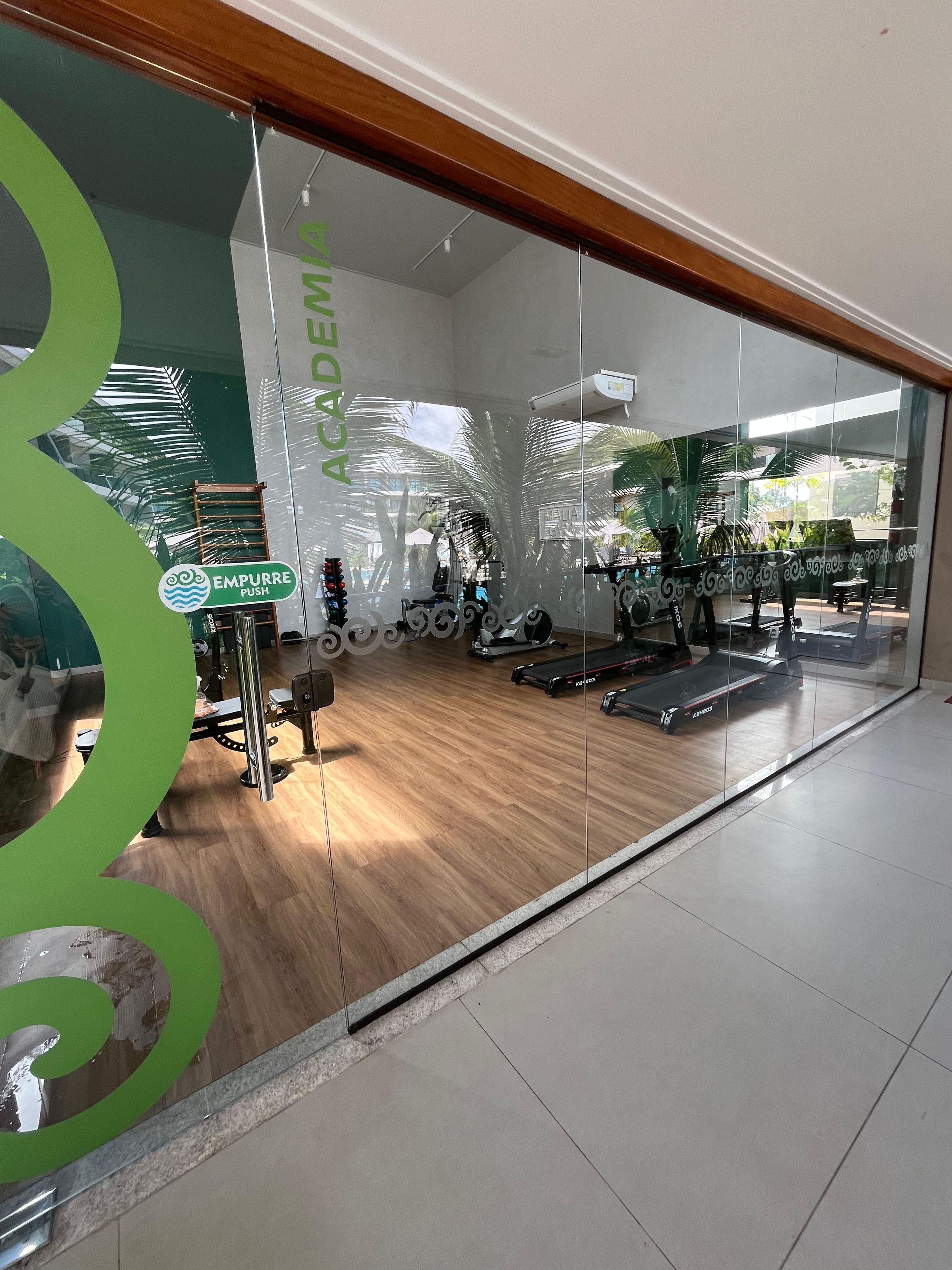 A gym with exercise equipment, a wooden floor, and a glass wall with a green logo.