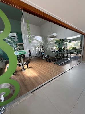 Une salle de sport équipée de matériel d’exercice, dotée d’un plancher en bois et d’un mur en verre orné d’un logo vert.