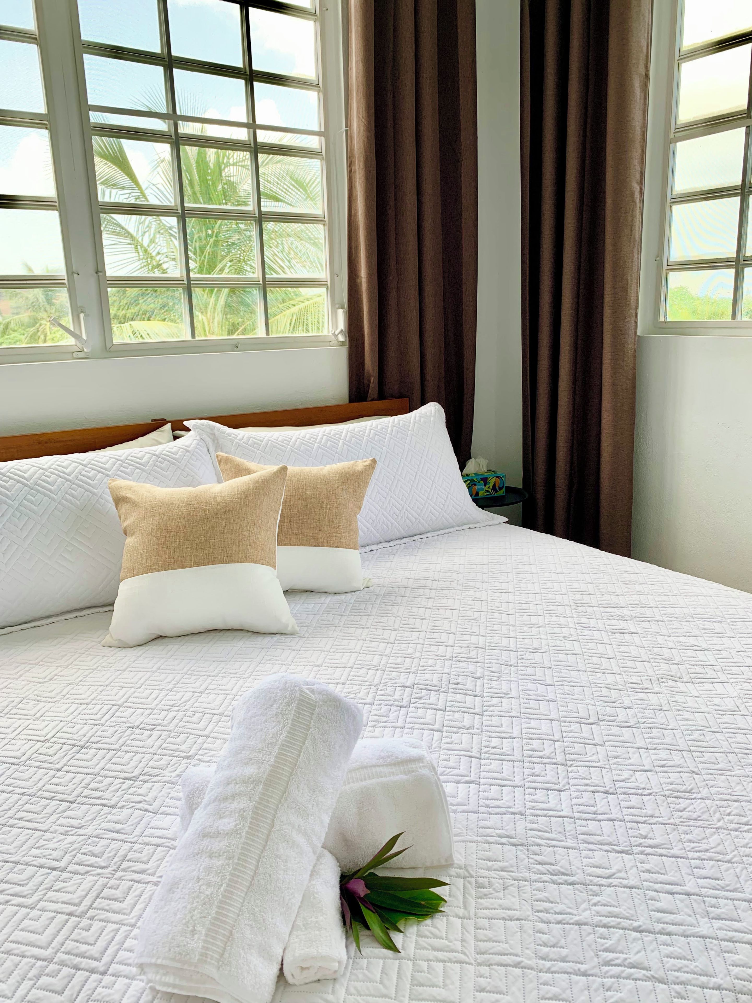 A neatly made bed with a quilted white bedspread, two beige pillows, and a rolled white towel with a green leaf decoration.