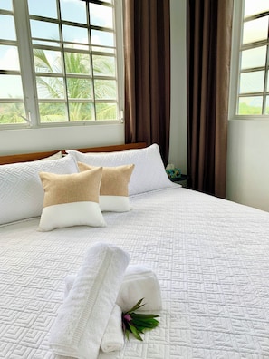 Een net opgemaakt bed met een gewatteerde witte spreidek, twee beige kussens en een opgerolde witte handdoek met een groene blad decoratie.