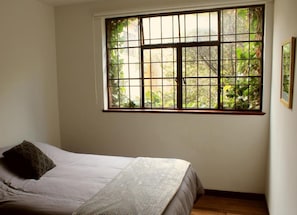 A bedroom with a bed, a window with bars, and a view of greenery outside. - Casa Muntú - La Candelaria (Bogotá)