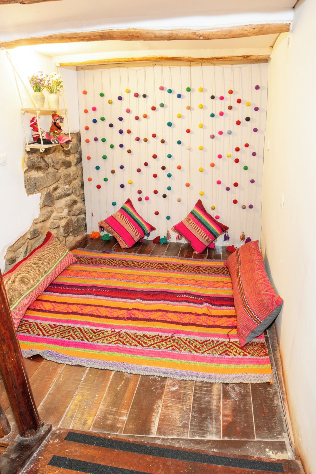 A cozy room with a stone wall, a colorful woven rug, and a wall hanging with colorful pom-poms.