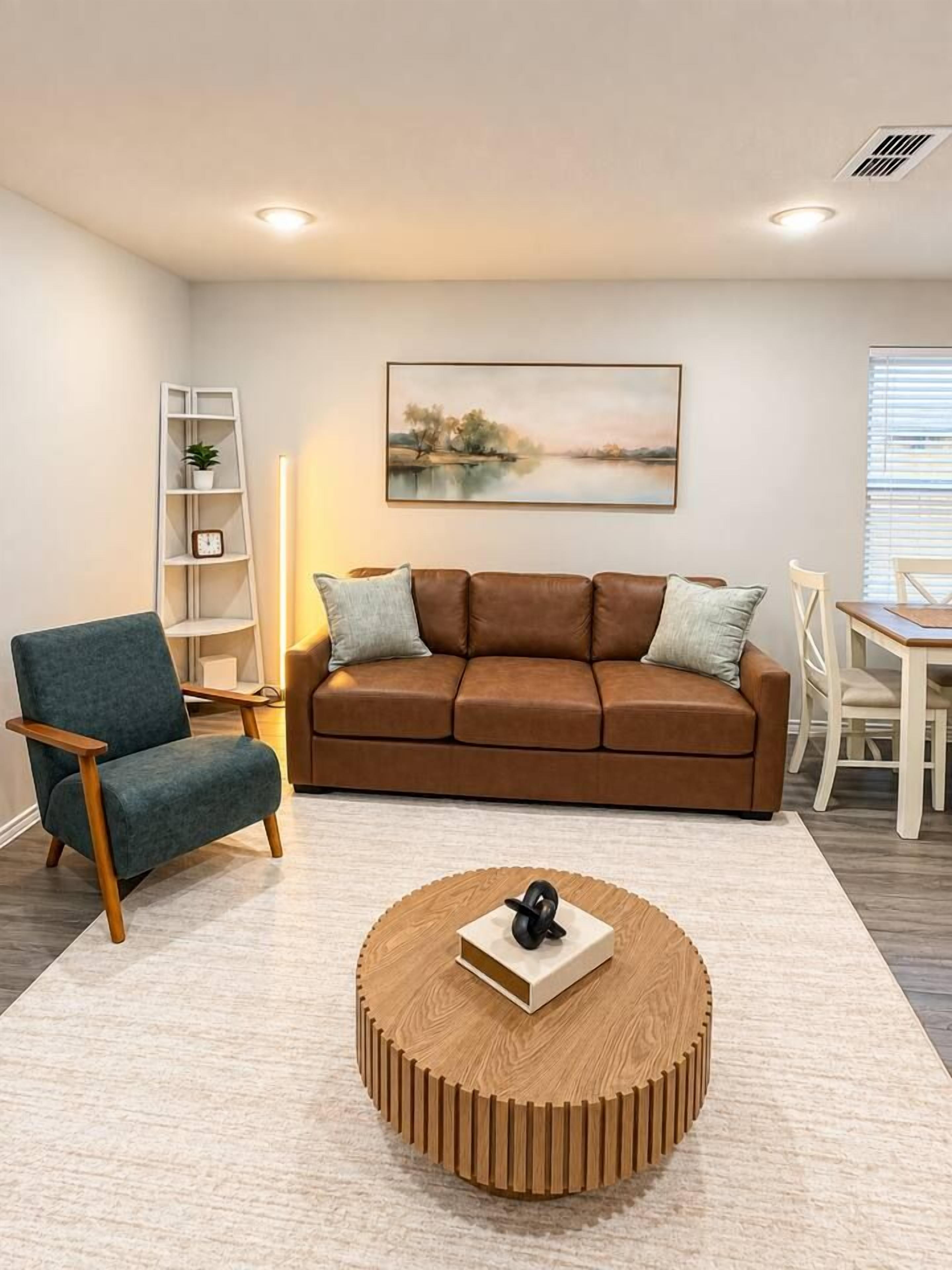 Een woonkamer met een bruine bank, een blauwe fauteuil, een houten salontafel, een schilderij aan de muur en een witte boekenkast.