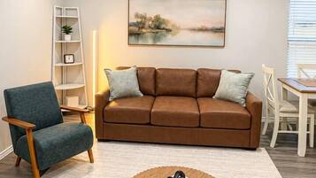 Een woonkamer met een bruine bank, een blauwe fauteuil, een houten salontafel, een schilderij aan de muur en een witte boekenkast.