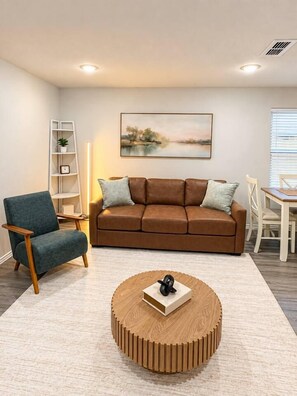 A living room with a brown sofa, a blue armchair, a wooden coffee table, a painting on the wall, and a white bookshelf. - Spacious 4BR Retreat | Near Dyess & Data Centers (Abilene)