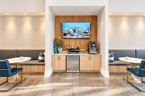 Eine moderne Hotelhalle mit einer zentralen Kaffeestation, einer Sitzecke und einem großen Fernseher, der eine Unterwasserwelt zeigt.