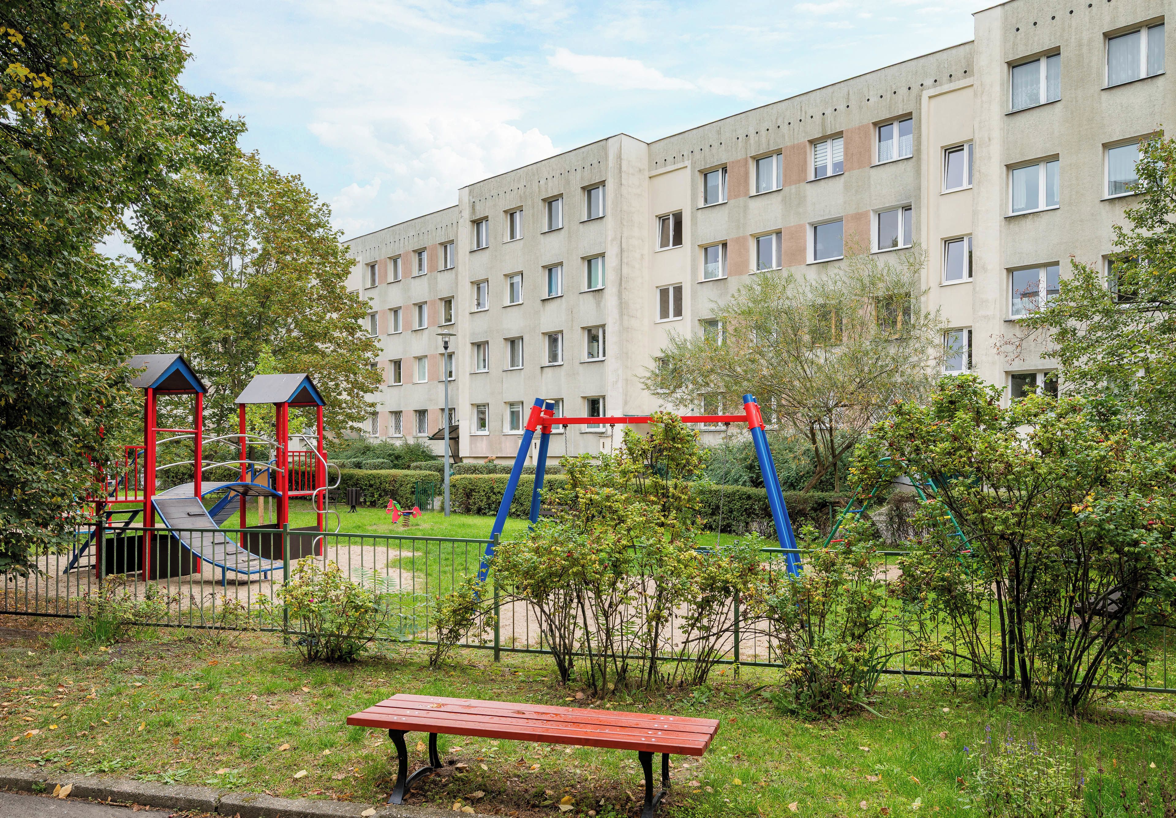 En legeplads med gynger og rutschebane, omkranset af et hegn, foran en bygning med mange vinduer.