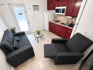 A compact living space with a sofa, a small table with a plant, a kitchenette with red cabinets, and a white refrigerator.