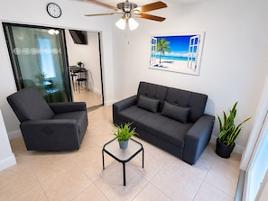 A modern living room with a sofa, armchair, and a small table with a plant. There is a ceiling fan, a clock, and a painting of a beach scene on the wall.