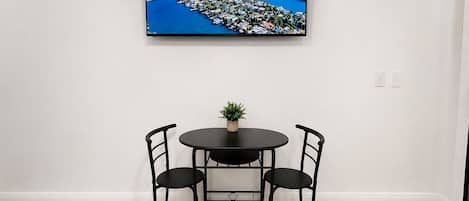 A dining area with a black table and chairs, a small potted plant, and a television mounted on the wall displaying a Miami advertisement.