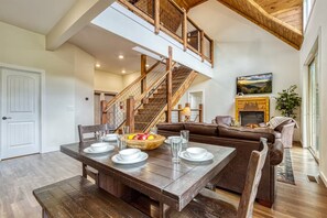 A dining area with a wooden table set for four, a staircase with wooden railings, and a living room with a fireplace. - Close to Pkwy~Htd Pool~View~Hot Tub~Arcade~Fam Fav (Sevierville)