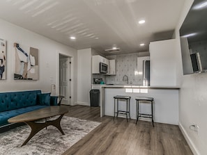 A modern kitchen with a central island, bar stools, and a blue sofa in a living area. - Remodeled Downtown 1Bed  #8 (Oklahoma City)