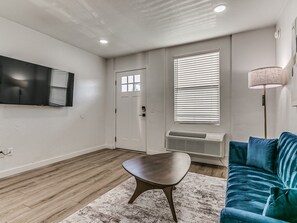 A modern living room with a flat-screen TV mounted on the wall, a door, a window with blinds, a standing lamp, a blue sofa, and a small wooden table. - Remodeled Downtown 1Bed  #8 (Oklahoma City)