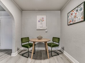 Et værelse med et rundt træbord, to grønne polstrede stole, et billede i ramme på væggen og en hvid dør i baggrunden.