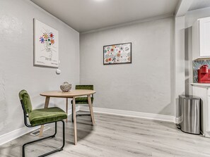 A room with a round wooden table, two green upholstered chairs, a small trash bin, and two framed artworks on the wall. - Stylish Midtown OKC Condo (Oklahoma City)