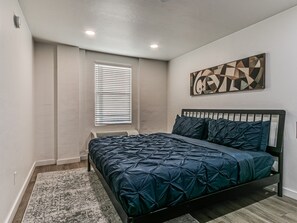 Ein Schlafzimmer mit einem Bett, einem Fenster mit Jalousien und einem an der Wand befestigten, abstrakten Kunstwerk.