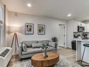 A modern living room with a grey sofa, a wooden coffee table, and a floor lamp. There is a kitchen area with white cabinets and a microwave. - Remodeled Urban Apartment 1 BR #1 (Oklahoma City)