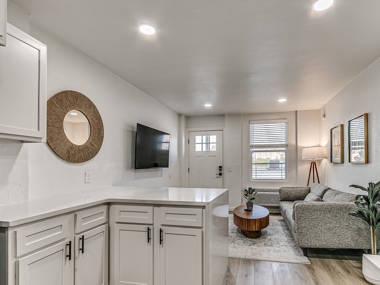 A modern kitchen with white cabinetry, a round mirror, a flat-screen TV, and a dining area with a round table and a sofa.
