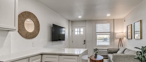 A modern kitchen with white cabinetry, a round mirror, a flat-screen TV, and a dining area with a round table and a sofa.