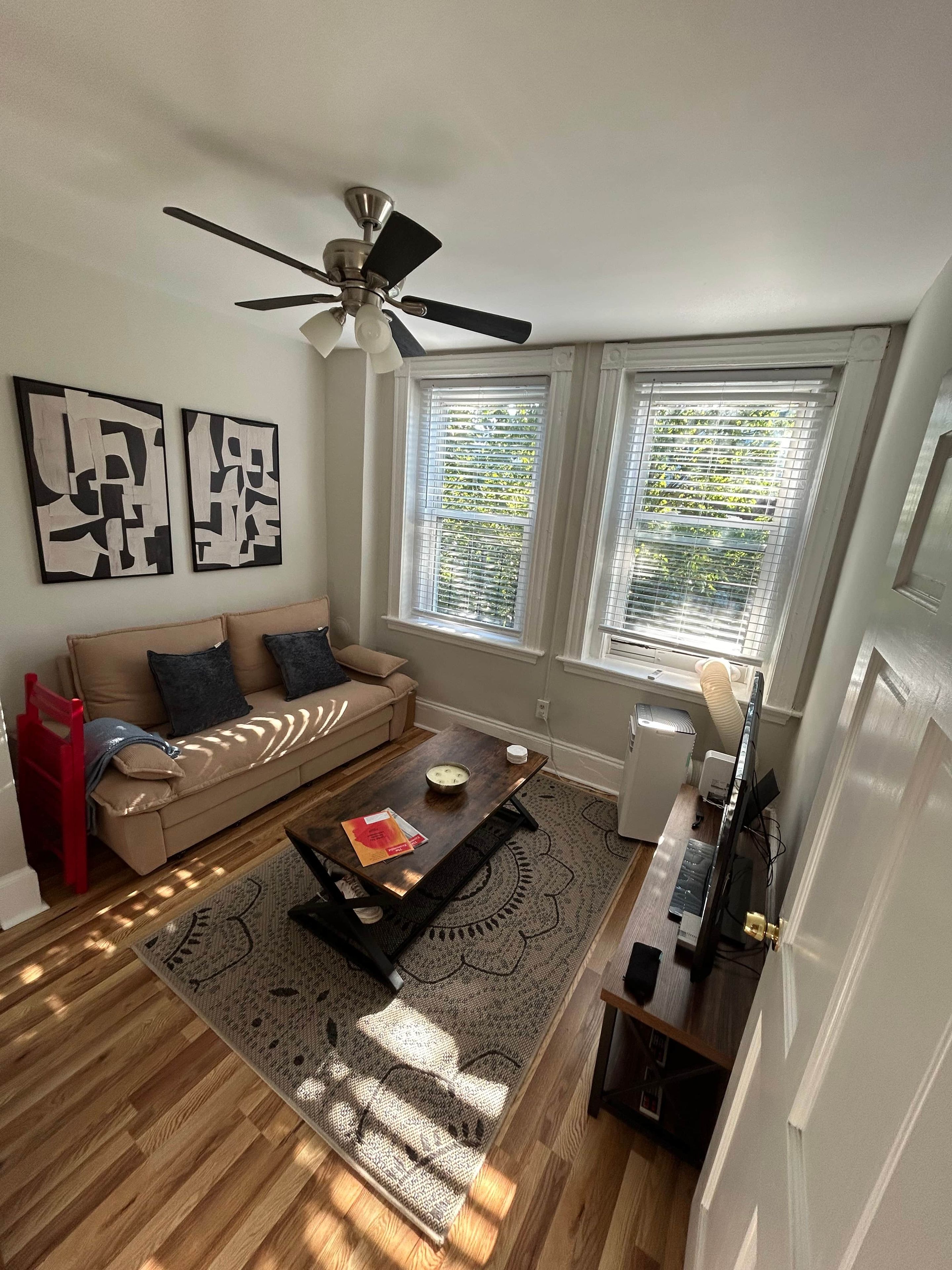 Un salon avec un ventilateur de plafond, un canapé, une table basse et deux fenêtres équipées de stores.