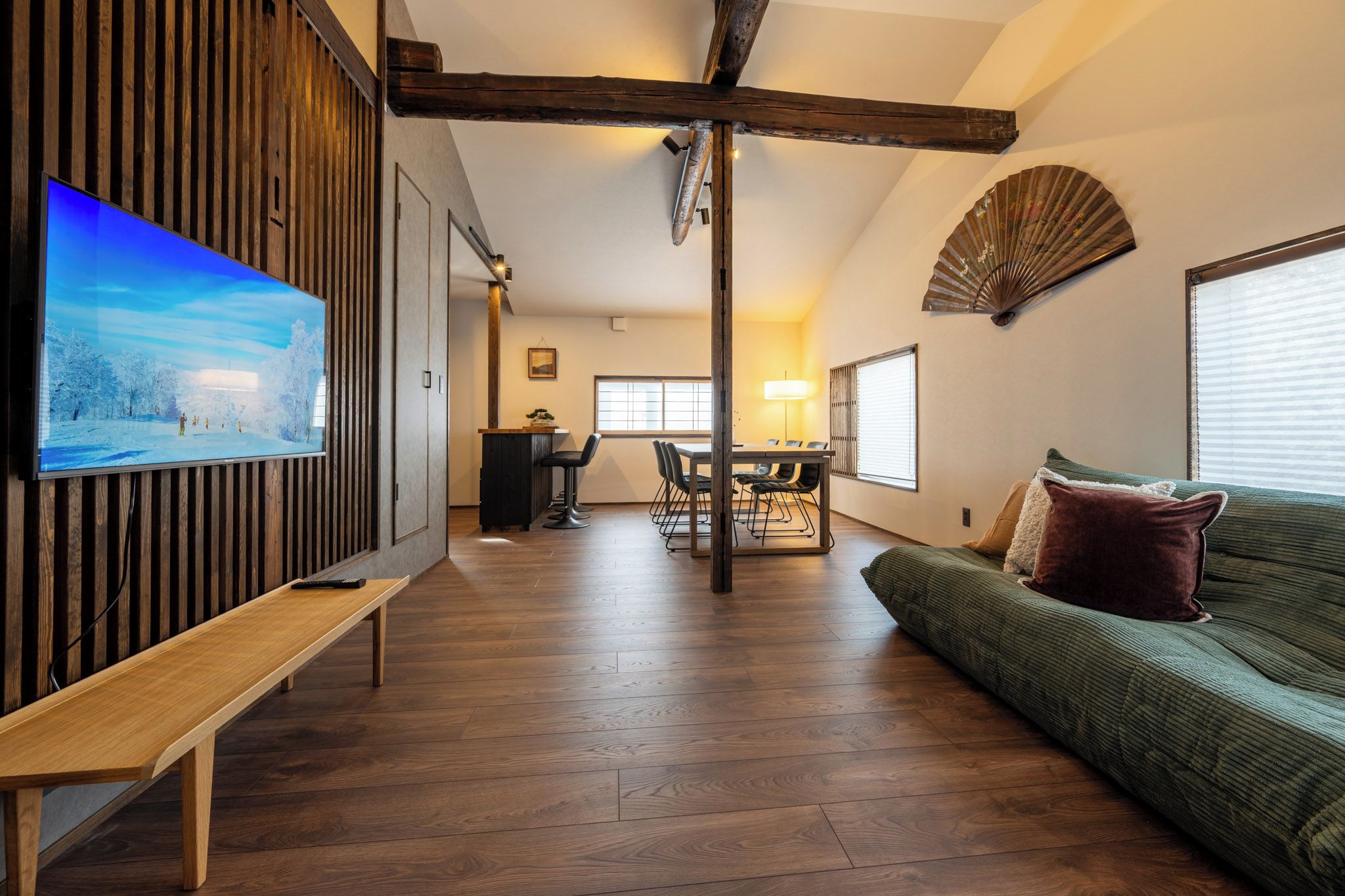 Uma sala de estar moderna com piso de madeira, um sofá verde, um banco de madeira, uma mesa de jantar com cadeiras e uma TV fixada em um painel de madeira.