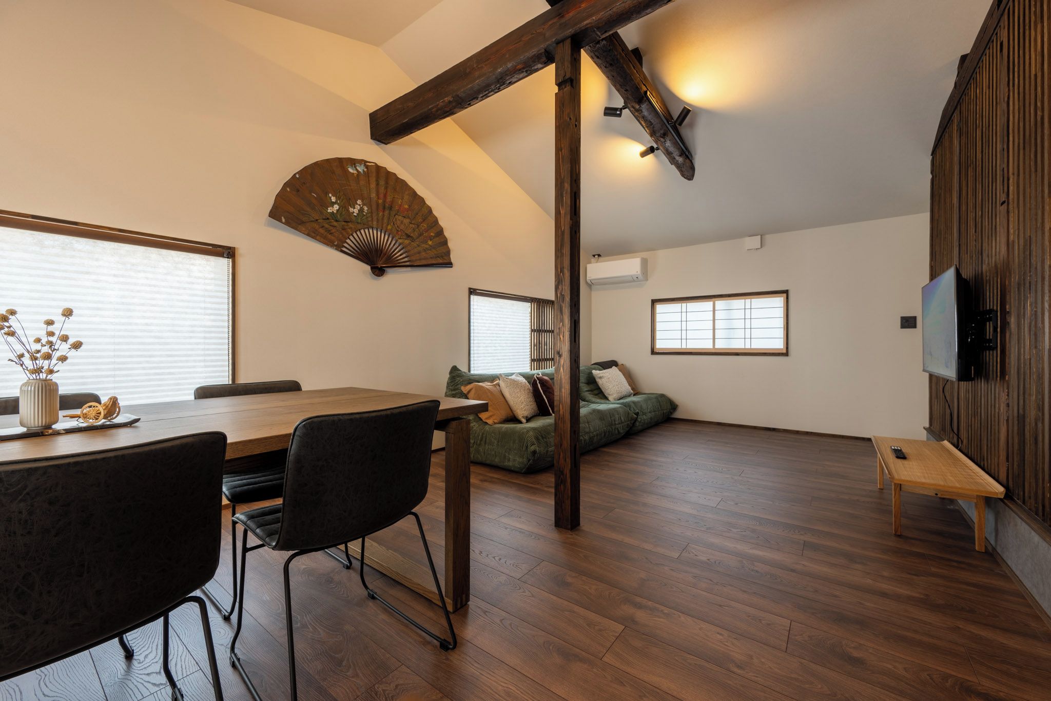 Um quarto amplo com piso de madeira, uma mesa de jantar com cadeiras, um sofá e uma televisão fixada na parede.