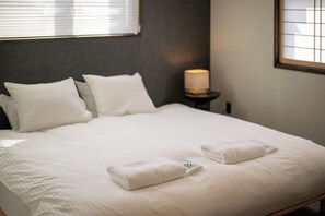 A neatly made bed with white linens and two folded towels, a bedside lamp, and a window with blinds. - Maru  (Shimotakai-gun)