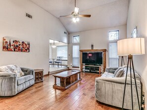 Ein geräumiges Wohnzimmer mit Holzdielenboden, Deckenventilator, Kamin und Fernseher.