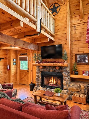 A log cabin interior with a stone fireplace, a mounted television, and a cozy seating area. - Cozy Cabin: Private Hot Tub, Fireplace & Mtn Views (Sevier County)