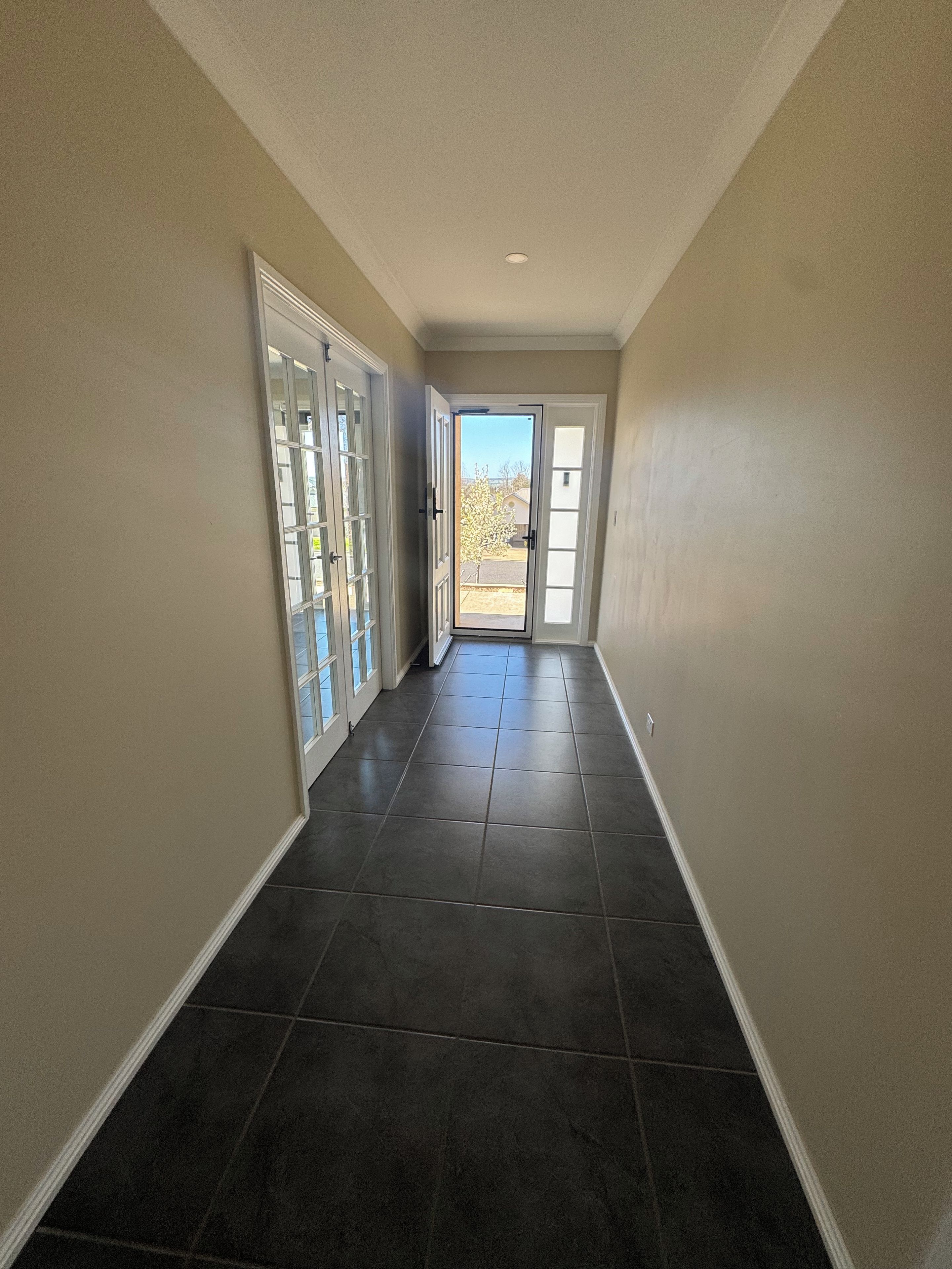 A hallway with dark tiled flooring, beige walls, and a door leading to a bright outdoor area.