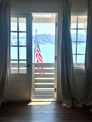 An open door leading to a balcony with an American flag, a view of the water, and a wooden floor.