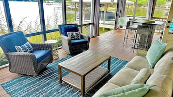 Een overdekt terras met rieten meubels, een houten tafel en uitzicht op een meer.