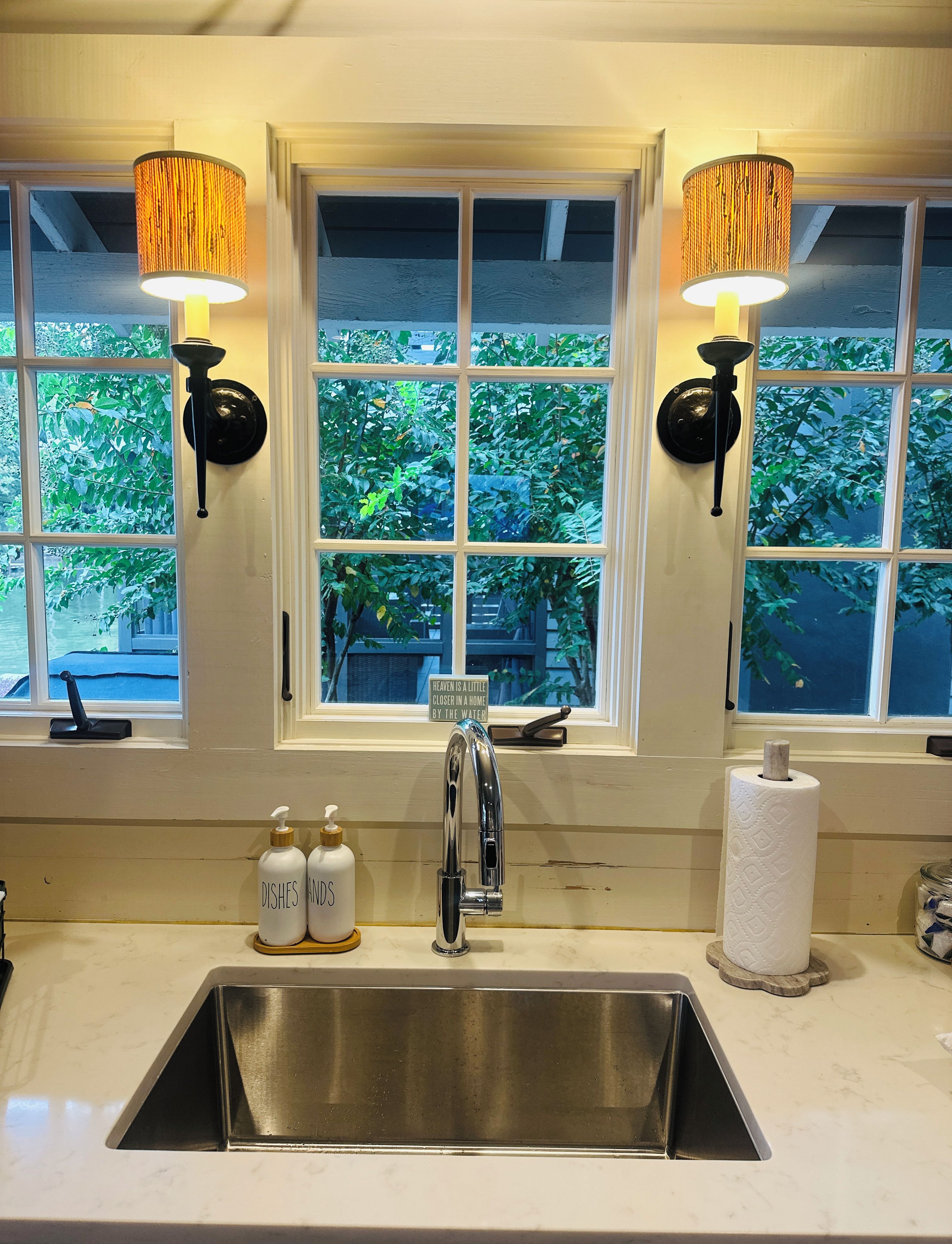 A kitchen with a stainless steel sink, two wall-mounted lamps, and a window with a view of greenery.